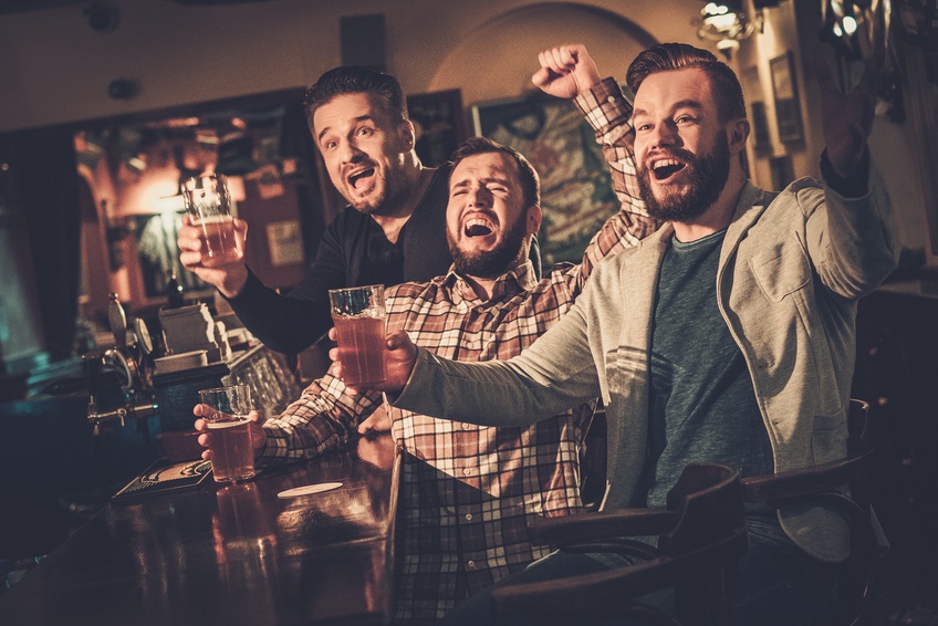 Cheerful old friends having fun watching a football game on TV and drinking draft beer at bar counter in pub.